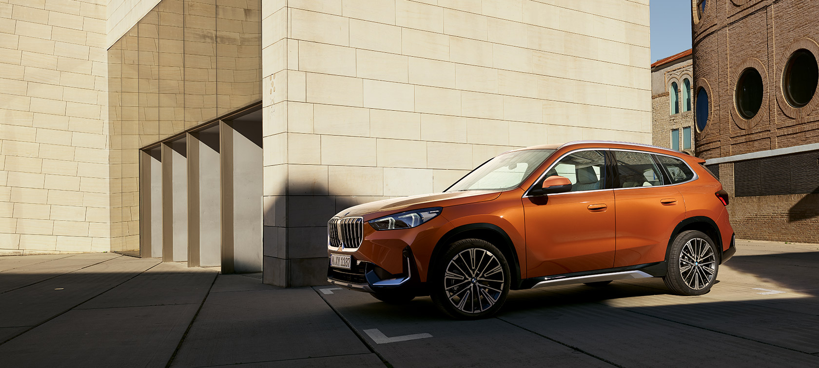 BMW U11 X1 xLine Utah Orange metalizado, vista de tres cuartos del frontal, parado delante de un edificio